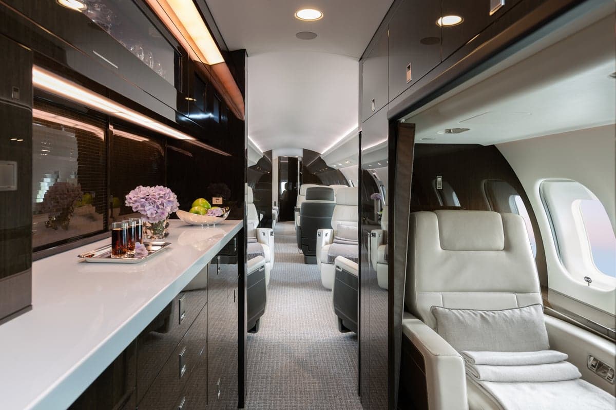 View down the aisle of the aircraft cabin with white leather seats and dark wood cabinets.