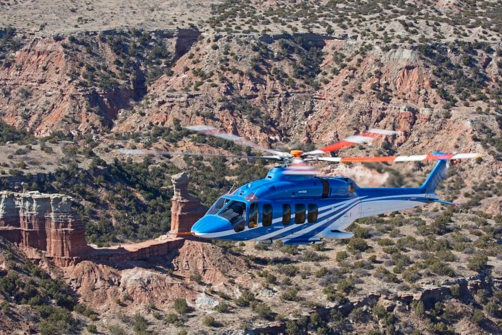 A blue and white Bell 525 helicopter flying over a canyon landscape.
