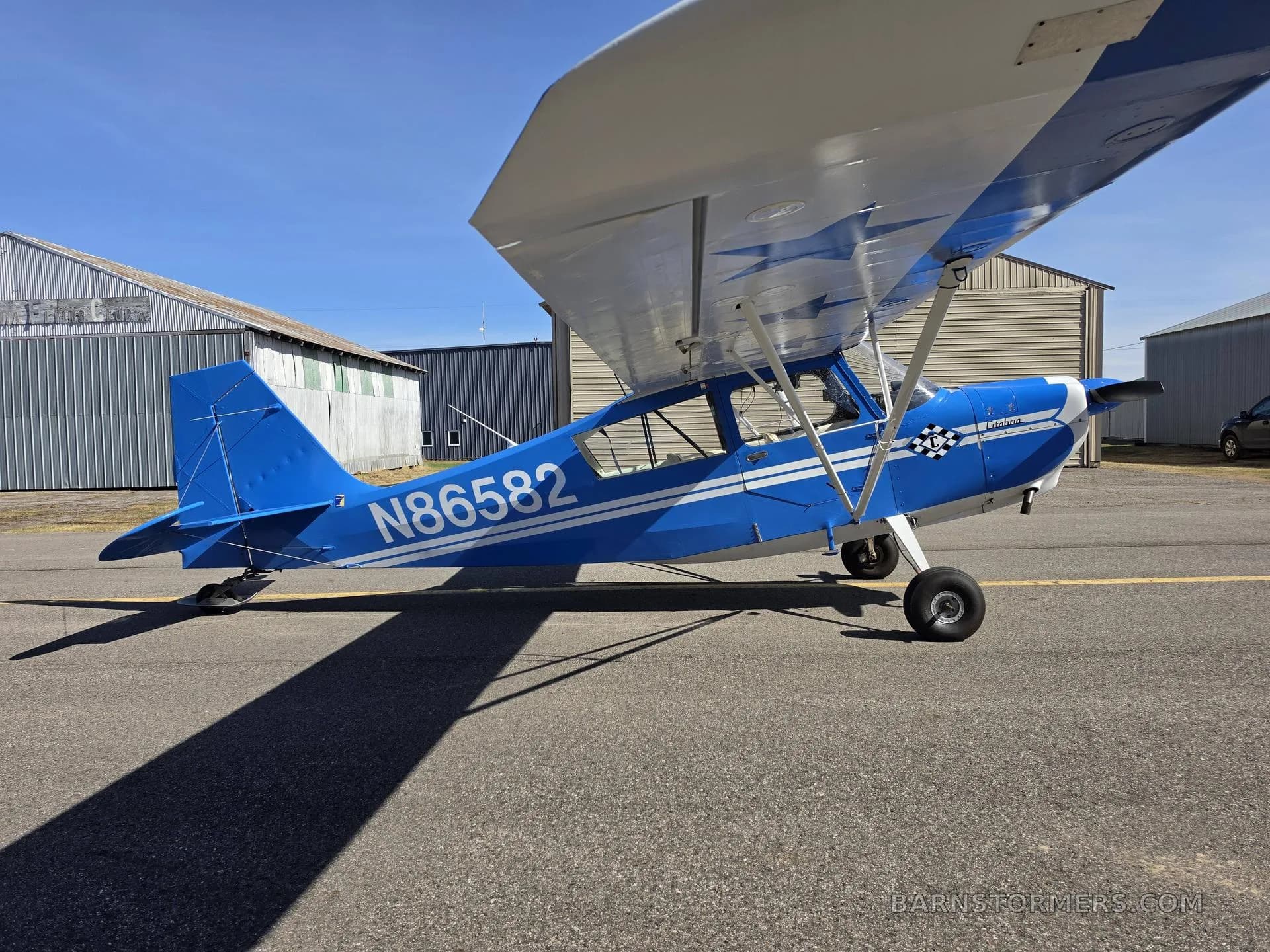 A blue and white single-engine propeller aircraft, tail number N86582, parked on an asphalt ramp with hangars in the background.