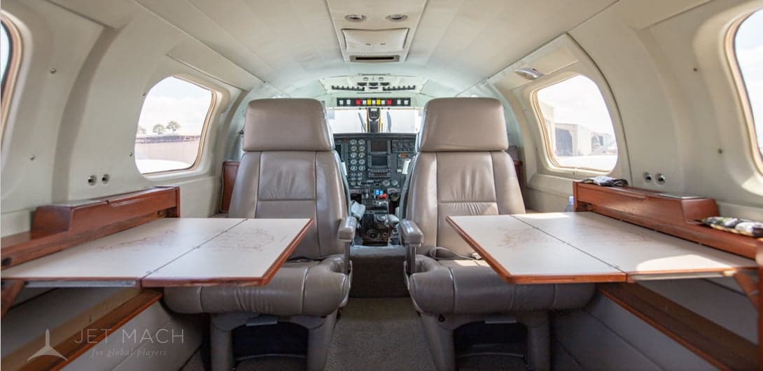 The aircraft cabin interior features beige leather seats, fold-out wooden tables, and a view into the cockpit.