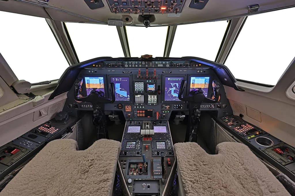 The cockpit of a private jet with multiple digital displays, control panels, and sheepskin seat covers.