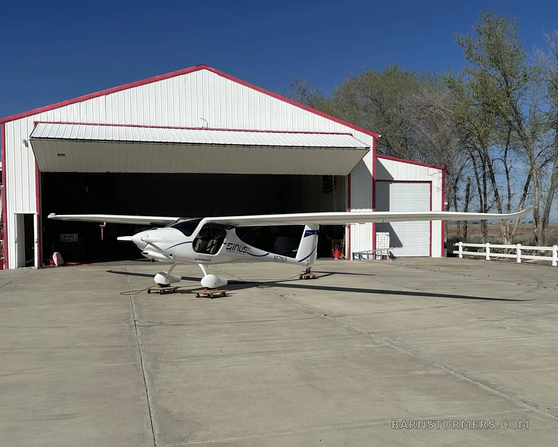 A white Pipistrel Sinus aircraft with blue accents is parked on an apron in front of a hangar on a sunny day.