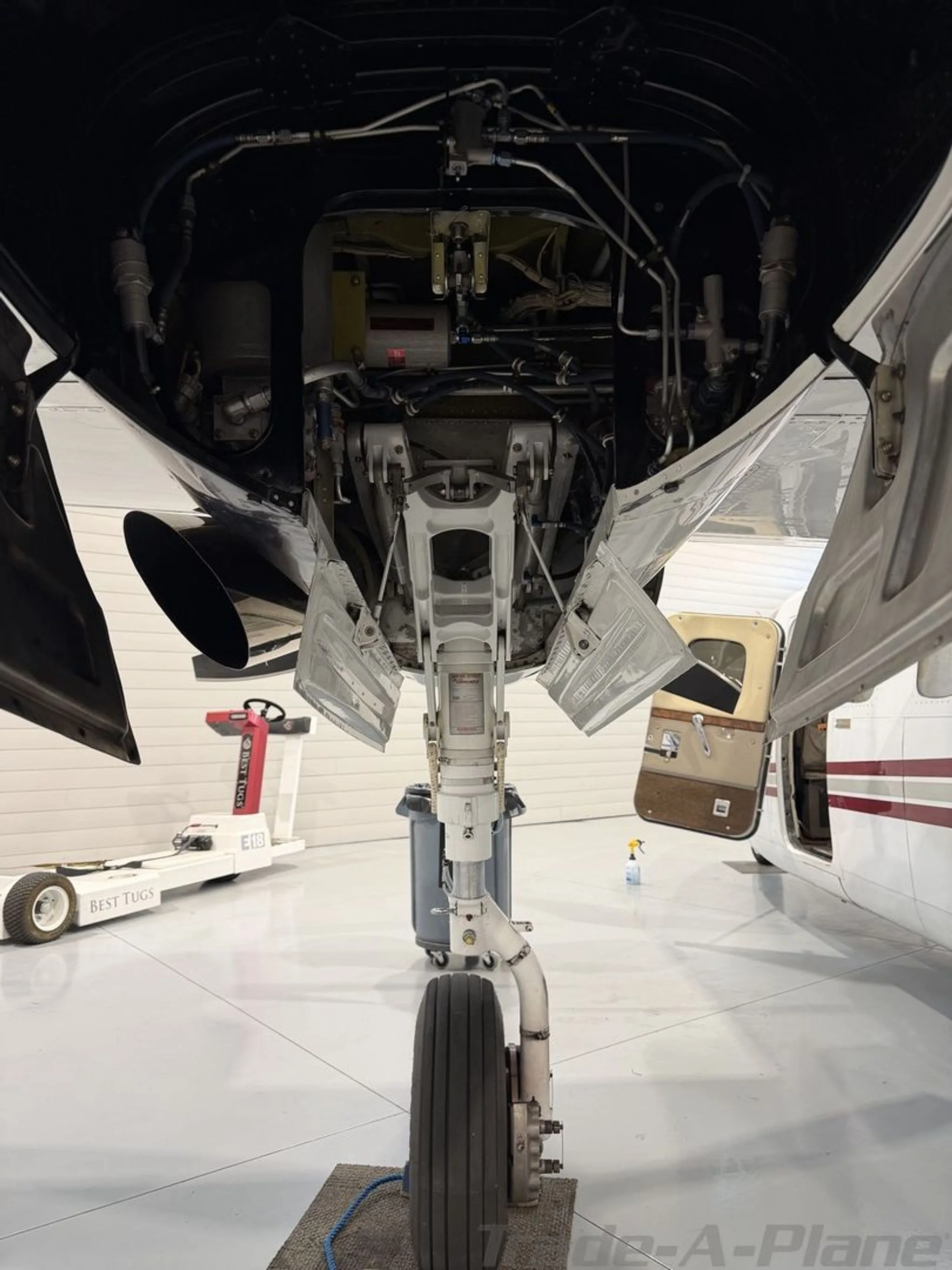 The nose landing gear and open access panels on the underside of a white aircraft in a hangar.