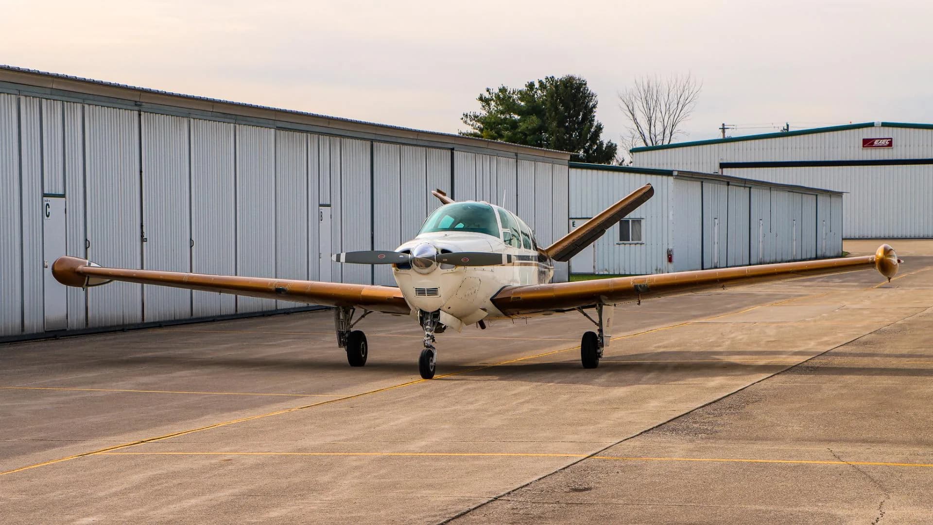Front three-quarter view of a white and gold Beechcraft Bonanza parked on an airfield.