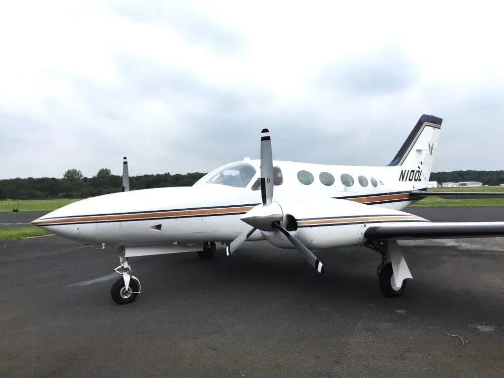 A white Cessna 421 Golden Eagle aircraft with blue and gold stripes parked on the tarmac under a cloudy sky.