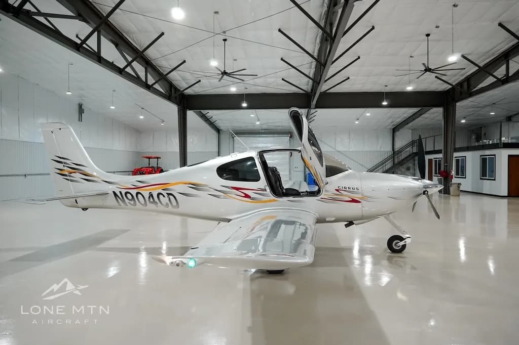 A silver Cirrus aircraft with red and yellow accent stripes, registration N904CD, parked in a hangar with its doors open.