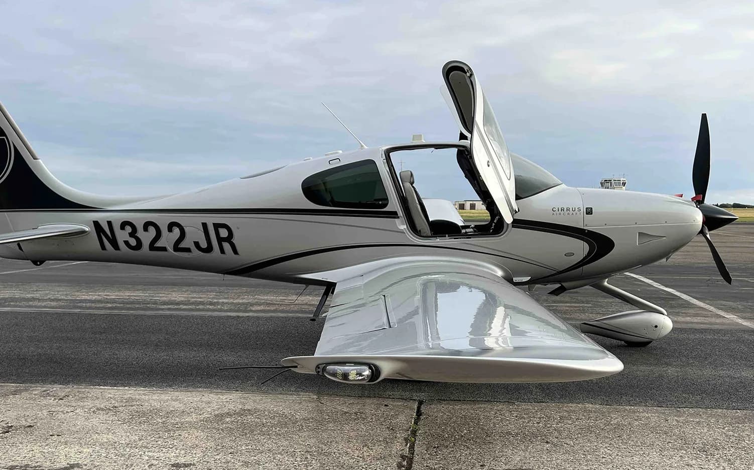 A silver Cirrus SR22 aircraft with registration N322JR and an open door is parked on an airfield.
