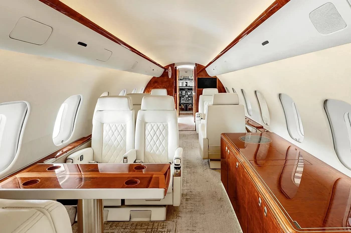 The spacious interior of a business jet features a dining table with cup holders, light-colored quilted seats, and rich wood cabinetry.