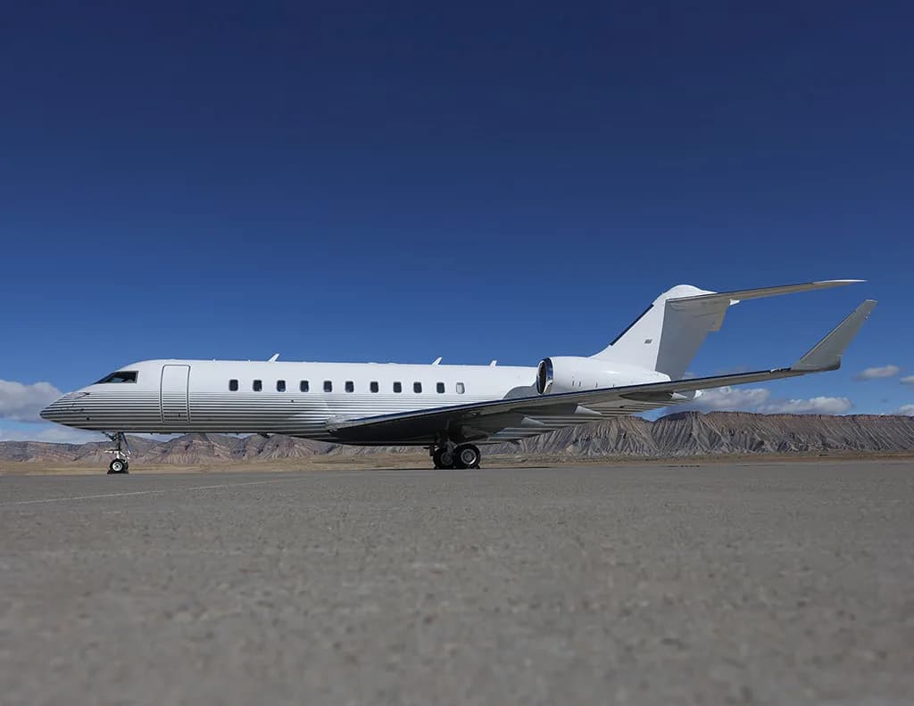 A white business jet with grey stripes on the fuselage is parked on an airfield under a clear blue sky.