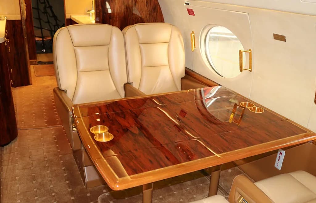 A close-up view of a private jet's polished wood dining table with gold cup holders and two tan leather seats.