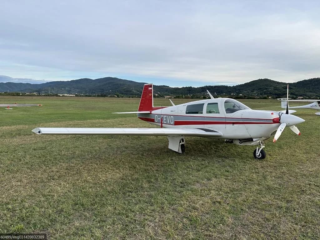 A white and red Mooney M20J aircraft (D-EEVO) parked on a grassy field with mountains in the background.