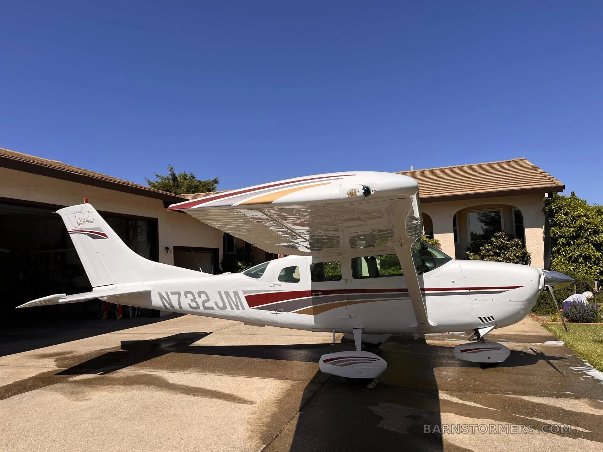 A white Cessna Skylane (N732JM) with red and gold stripes is parked on a wet driveway next to a house under a clear blue sky.