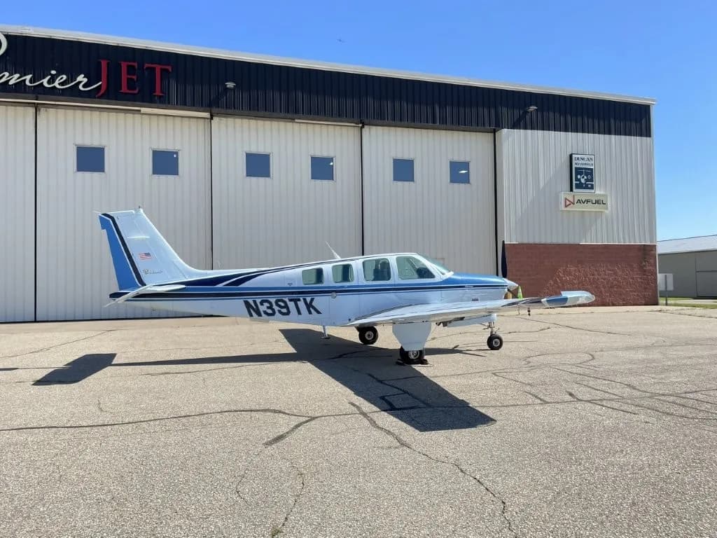A white and blue Beechcraft Bonanza N39TK parked on the tarmac in front of a hangar.