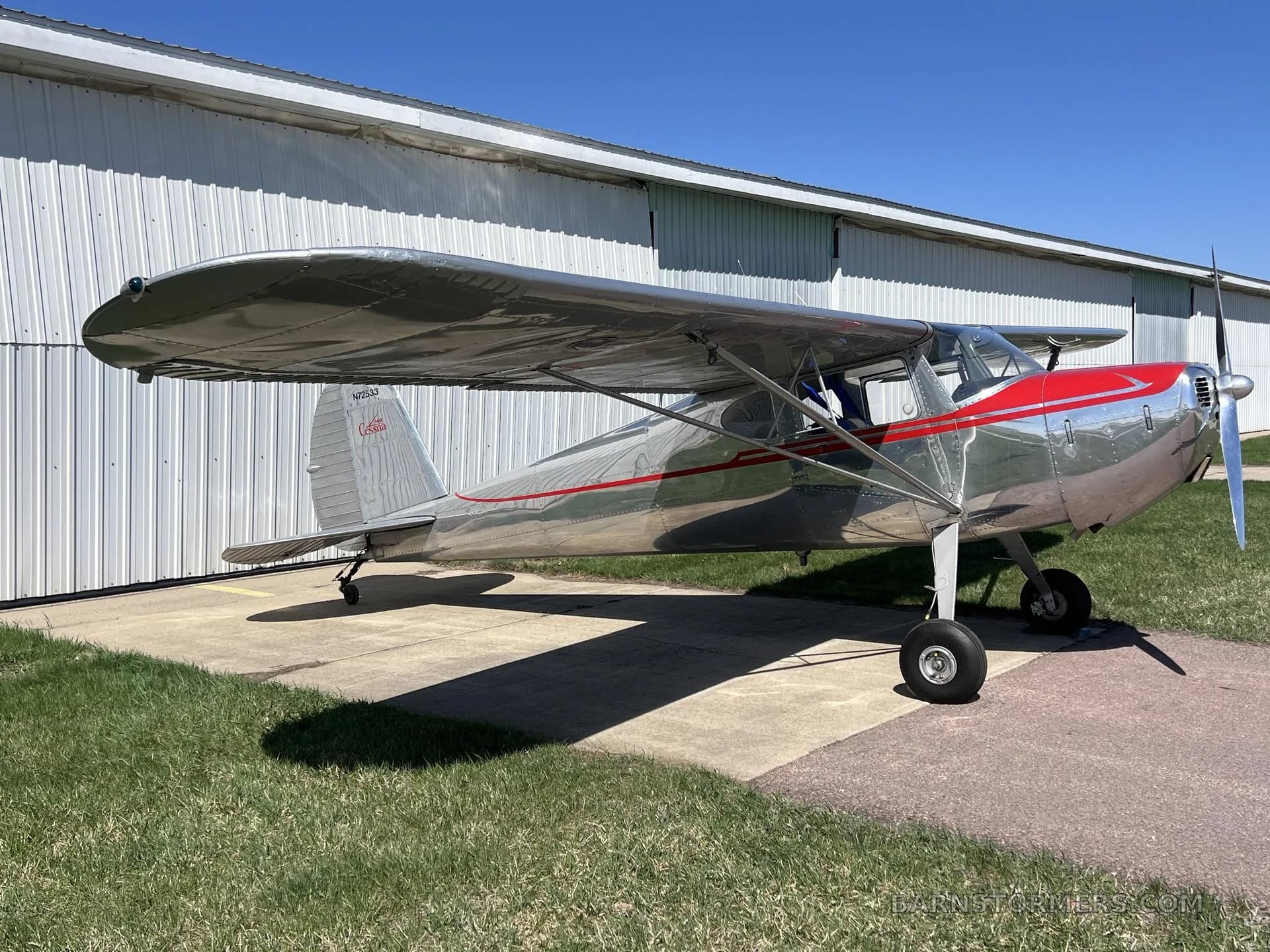 A silver Cessna 140 with red accents on the cowling and fuselage, parked on a concrete pad next to a white hangar under a clear blue sky.