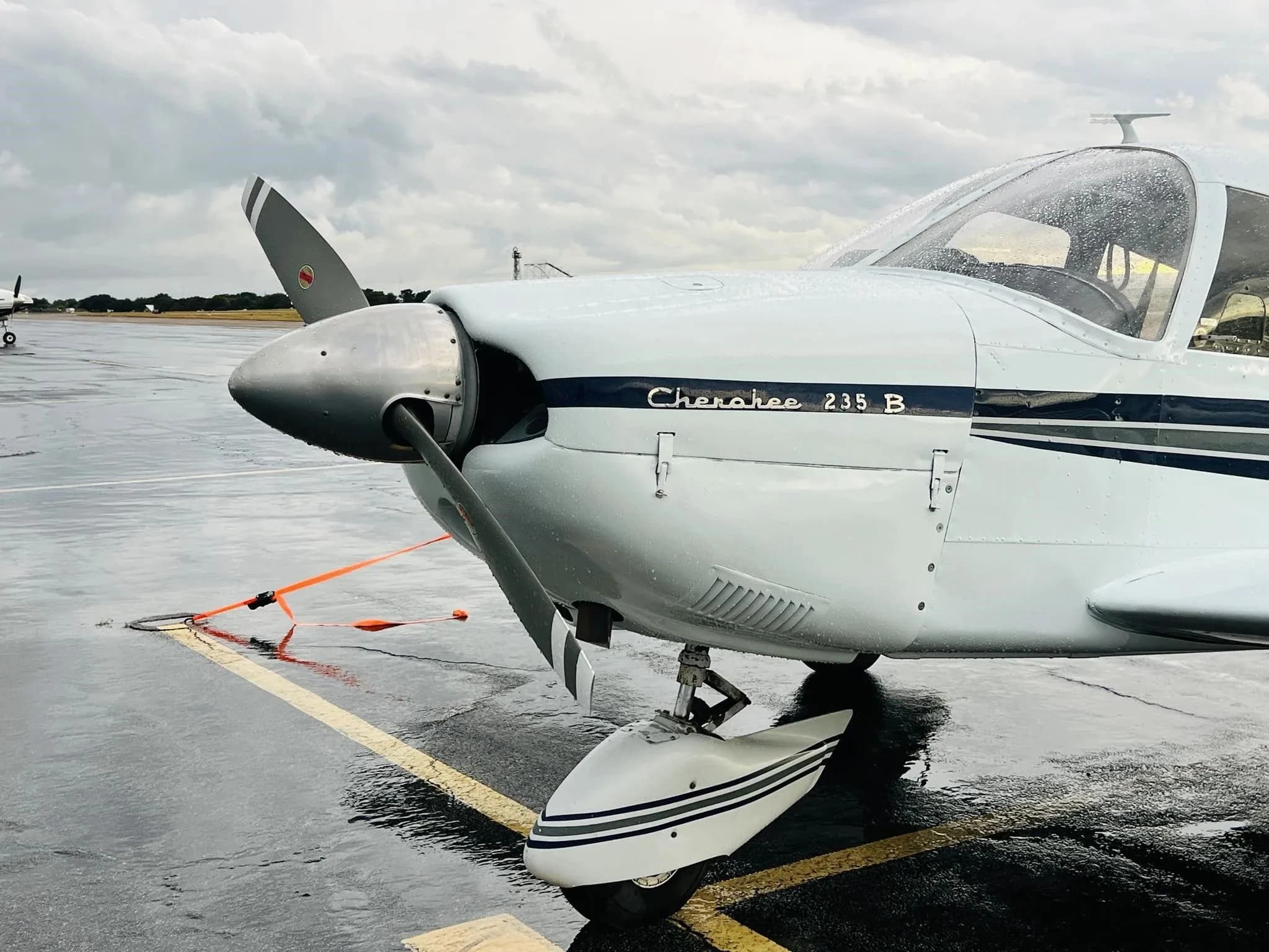 Close-up of the nose and propeller of a white Piper Cherokee 235 B on a wet tarmac.