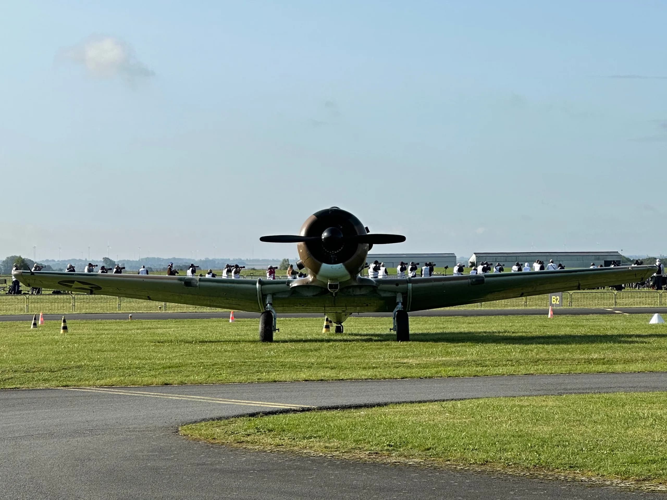 A North American T-6 Texan aircraft, F-AZAU, in camouflage paint, viewed head-on on an airfield with spectators in the background.