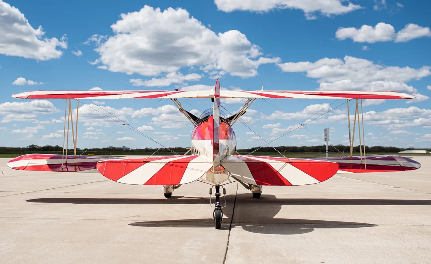 Rear view of a red and white biplane on the tarmac with a blue sky and clouds.