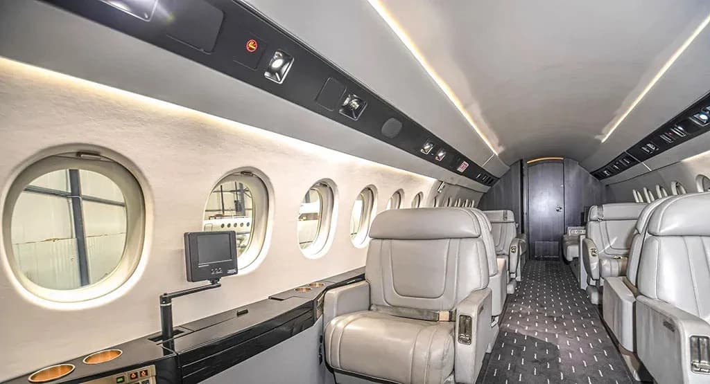 A side view of the Dassault Falcon 900EX cabin, showing light-colored seats, windows, and a small entertainment screen.
