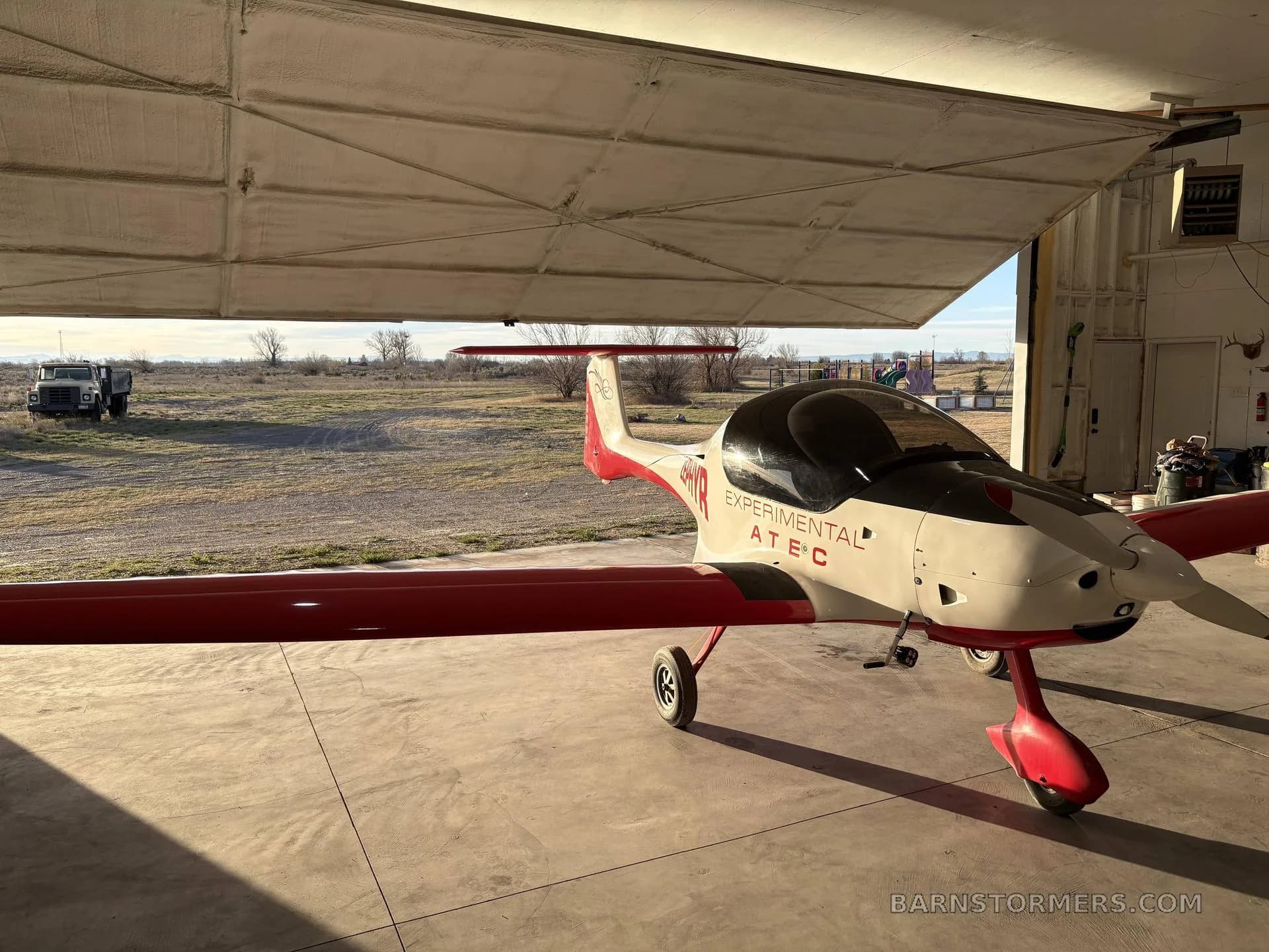 A white and red ATEC Zephyr experimental aircraft is parked inside a hangar, viewed from the front-left side.