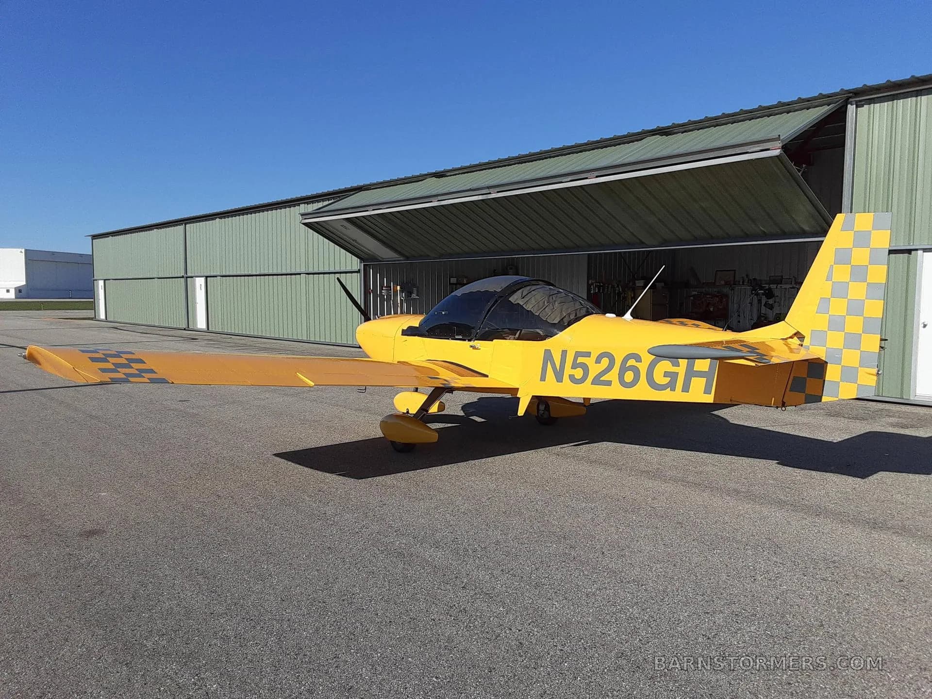 A bright yellow single-engine aircraft with registration N526GH and checkered tail/wing accents parked on an asphalt ramp in front of an open green hangar.