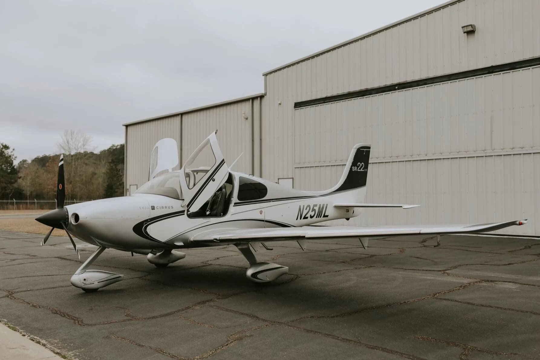 A silver Cirrus SR22 GTS aircraft with its gull-wing doors open, parked on an asphalt ramp in front of a hangar.