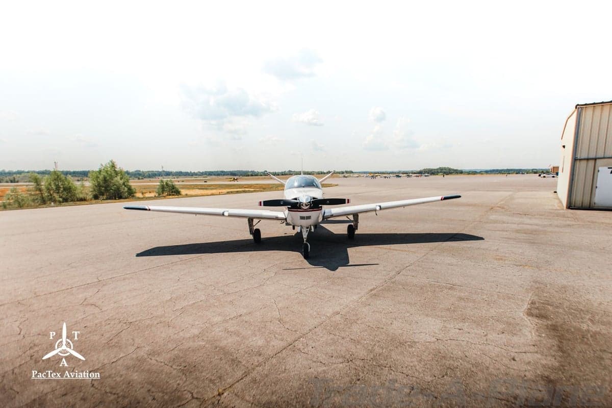 Frontal view of a white single-engine aircraft with red and black stripes parked on an airfield with a hangar in the background.