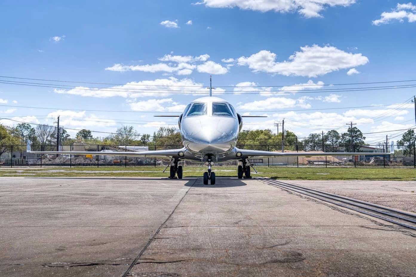 A head-on view of a silver business jet parked on an asphalt runway under a blue sky.