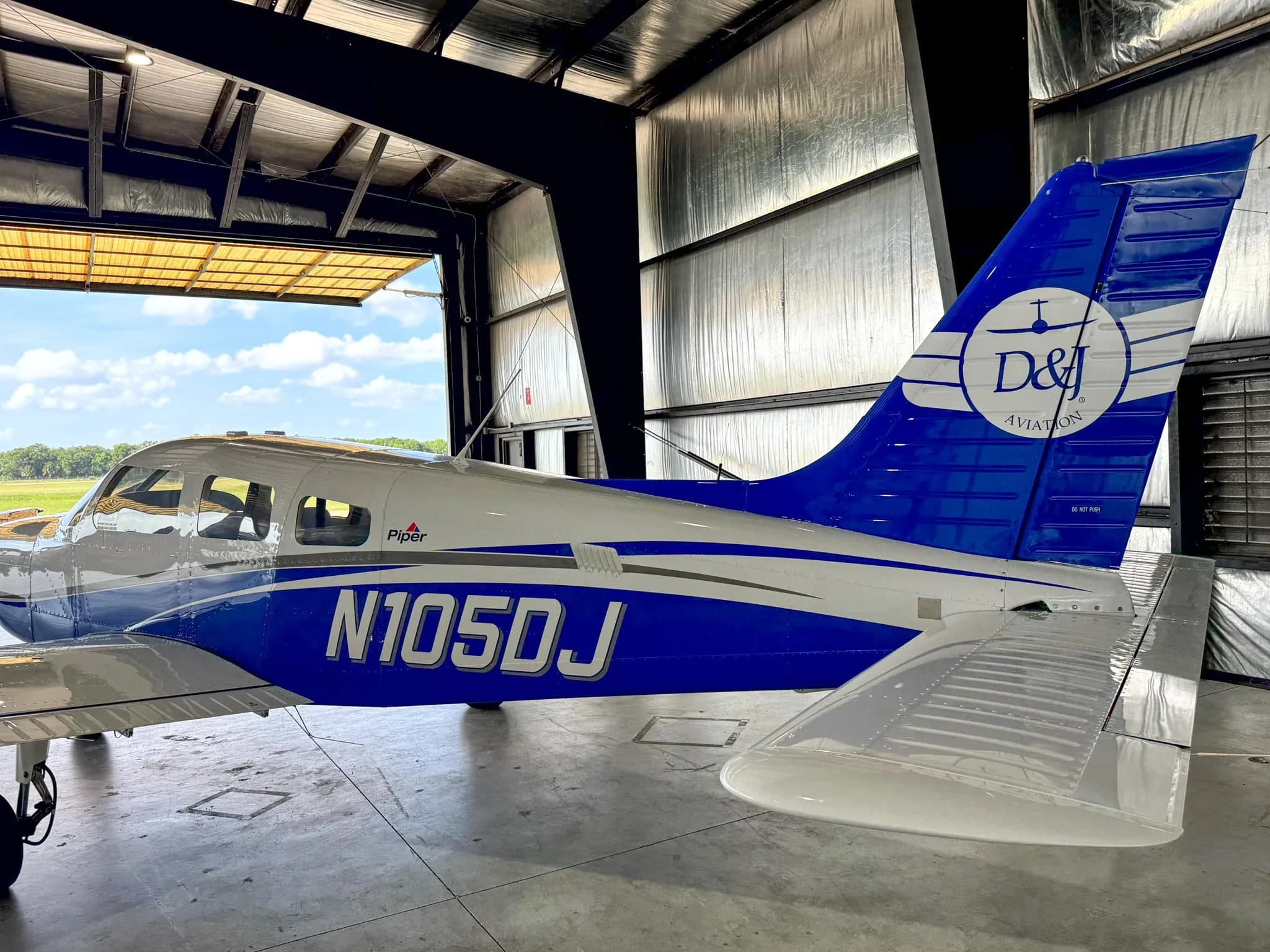 A side view of the white and blue Piper Archer TX, N105DJ, parked in a hangar with a view of the airfield outside.