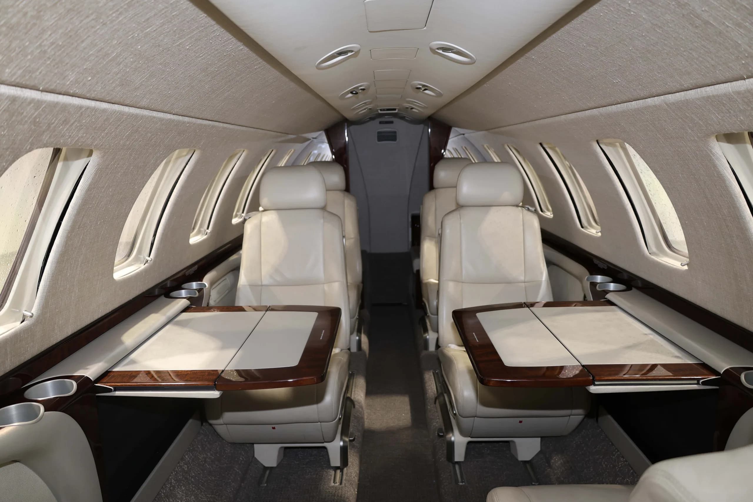 The interior cabin of a business jet featuring light beige leather seats and two fold-out wooden tables deployed.
