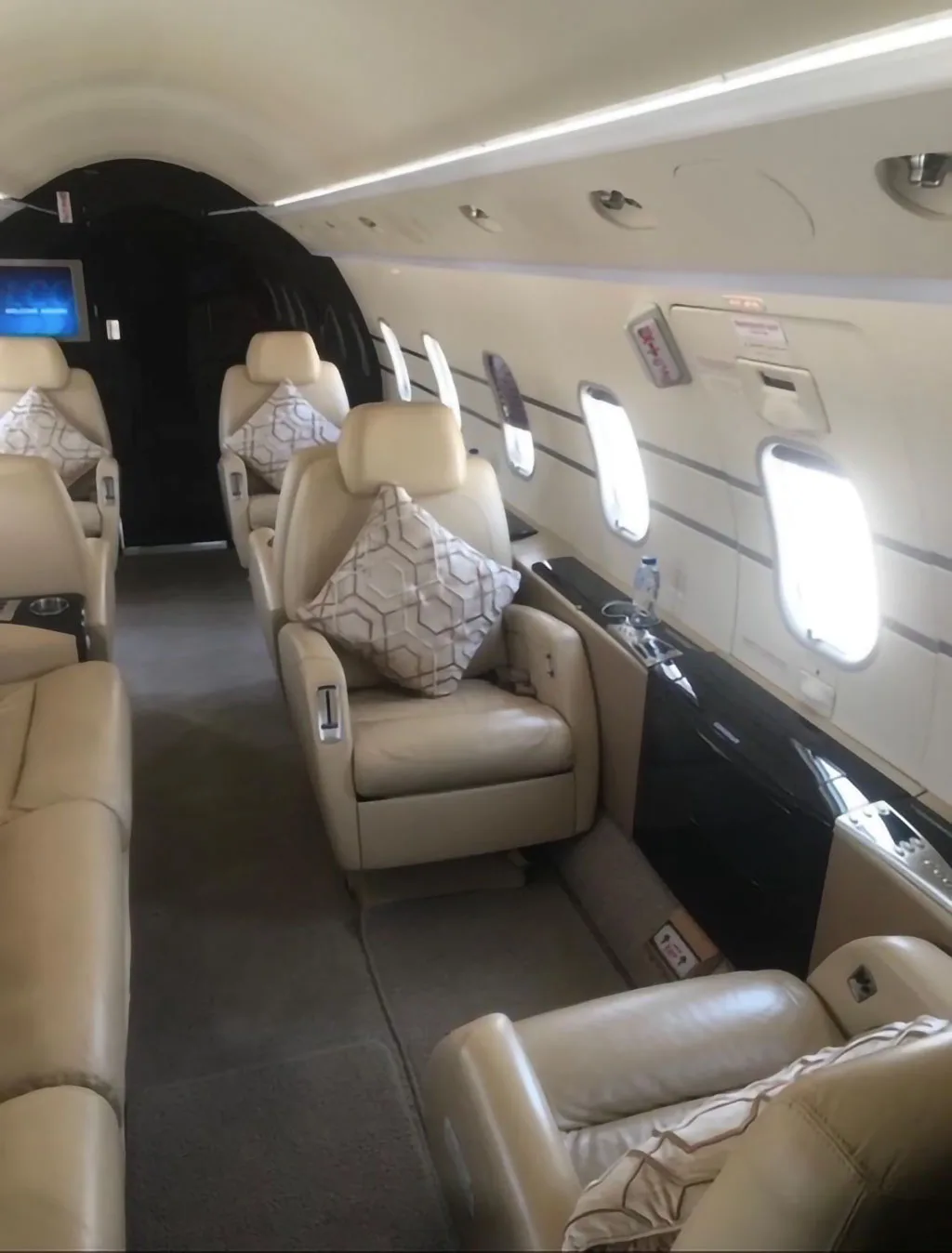 The interior of a private jet cabin features light beige leather seats with patterned pillows and rectangular windows.
