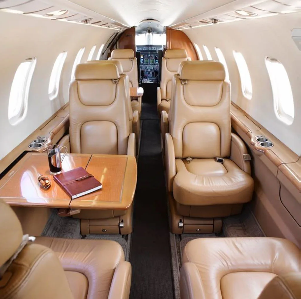 The aircraft cabin features tan leather seats and wooden tables, with a view towards the cockpit.