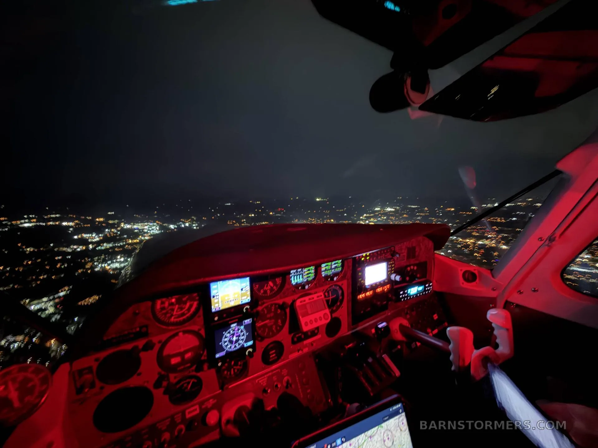 A night view from an aircraft cockpit, showing the illuminated instrument panel in red light and city lights sprawling below.