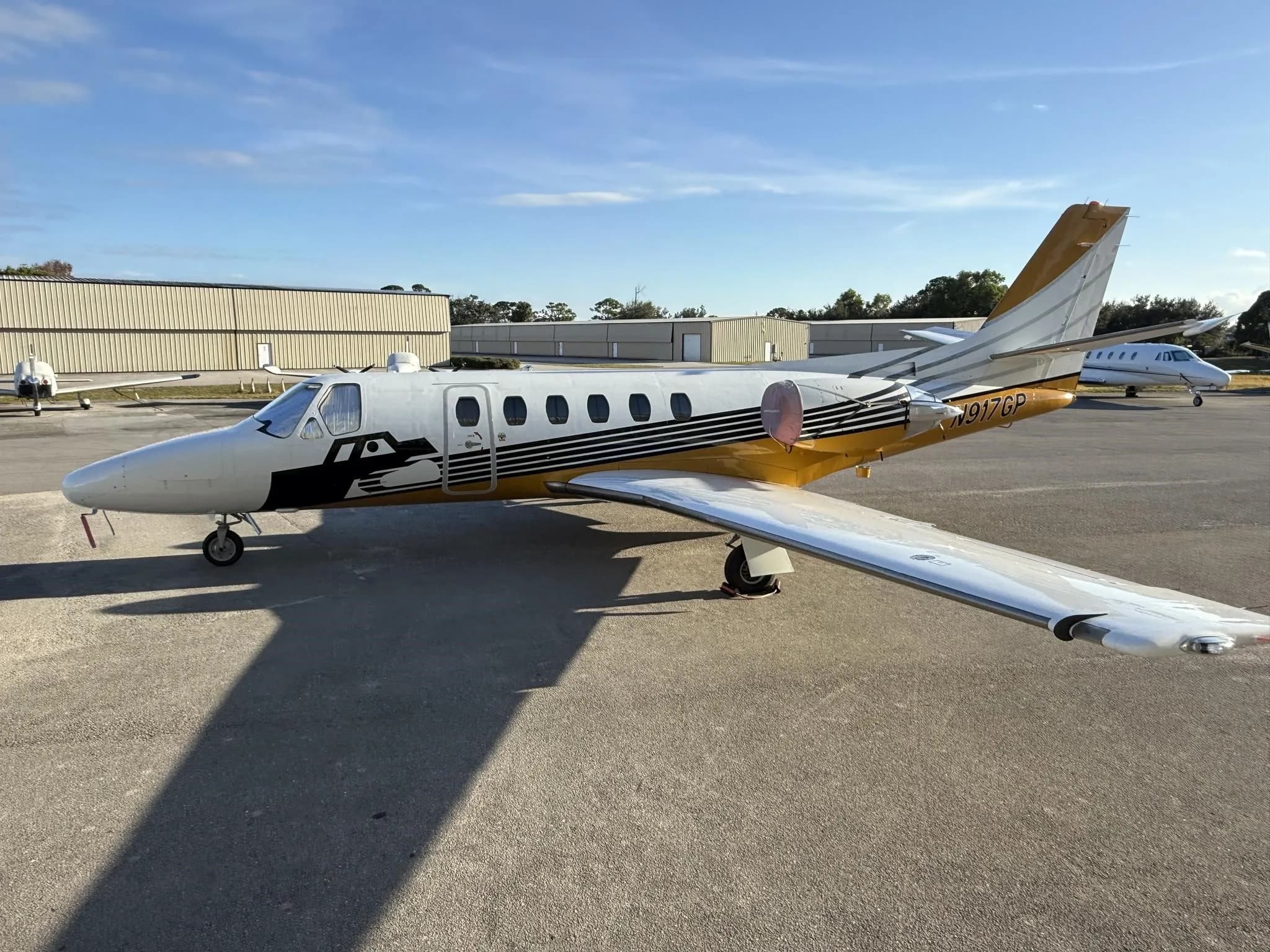A Cessna Citation jet, N917GP, with white and gold paint scheme, parked on an airfield under a blue sky.