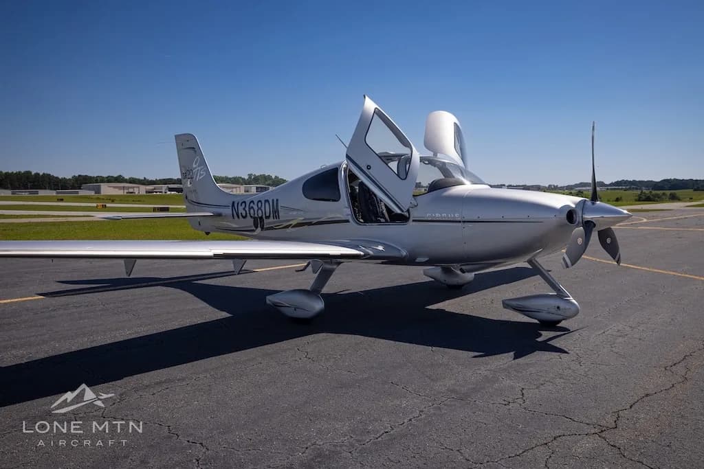A silver Cirrus SR22 Turbo aircraft (N368DM) with its doors open, parked on an asphalt runway under a clear blue sky.