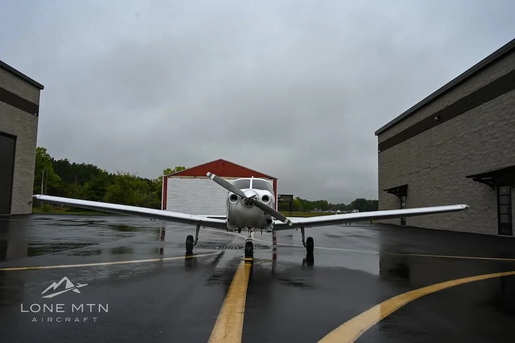 A white single-engine aircraft, N616AC, is viewed from the front on a wet tarmac with a red hangar in the background.