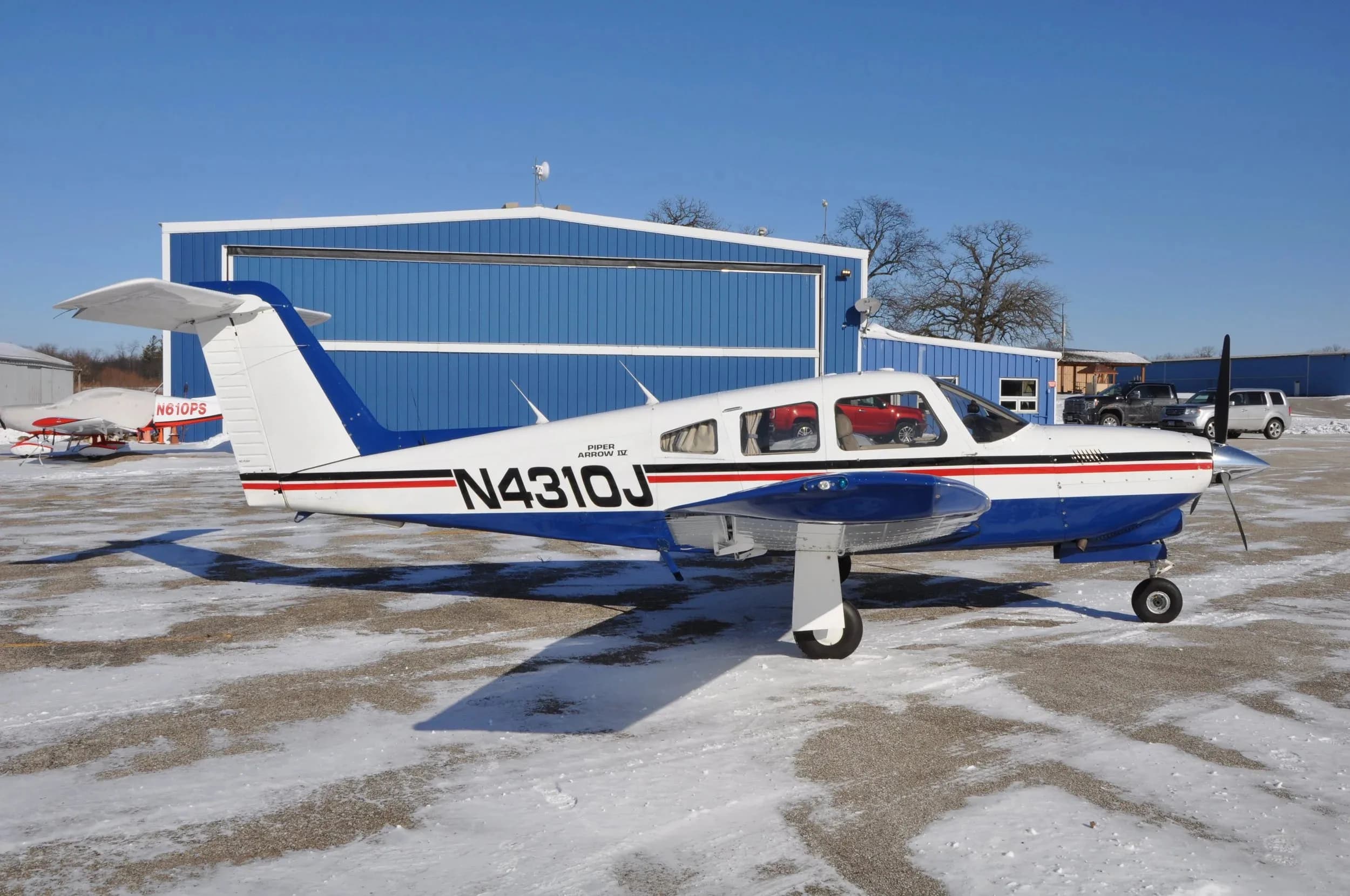 A white and blue Piper Arrow IV aircraft with registration N4310J parked on a snowy tarmac in front of a blue hangar.