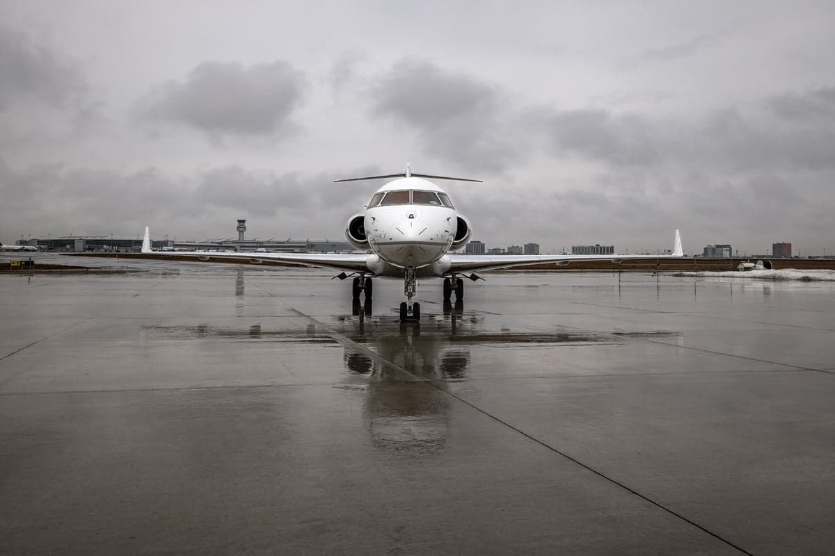 A white Bombardier Global Express stands on a wet tarmac under a cloudy sky.