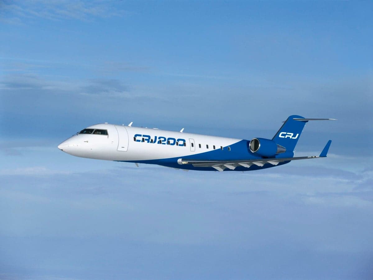 A white and blue Bombardier CRJ200 regional jet flying in a cloudy blue sky.