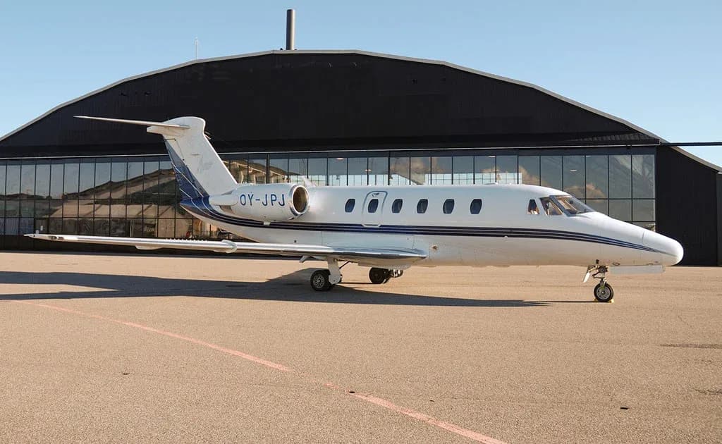 A white business jet with blue stripes parked on an airfield in front of a large black hangar.