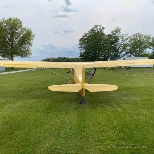 A yellow and orange single-engine high-wing aircraft is viewed from the rear, parked on a grassy field.