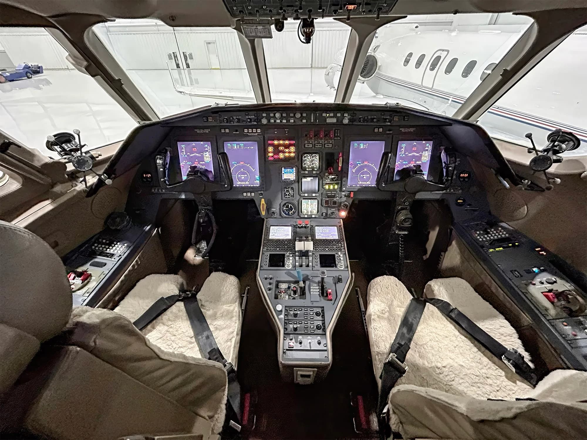 The cockpit of a Dassault Falcon 900EX, showing the instrument panel with multiple displays, controls, and two pilot seats with sheepskin covers.