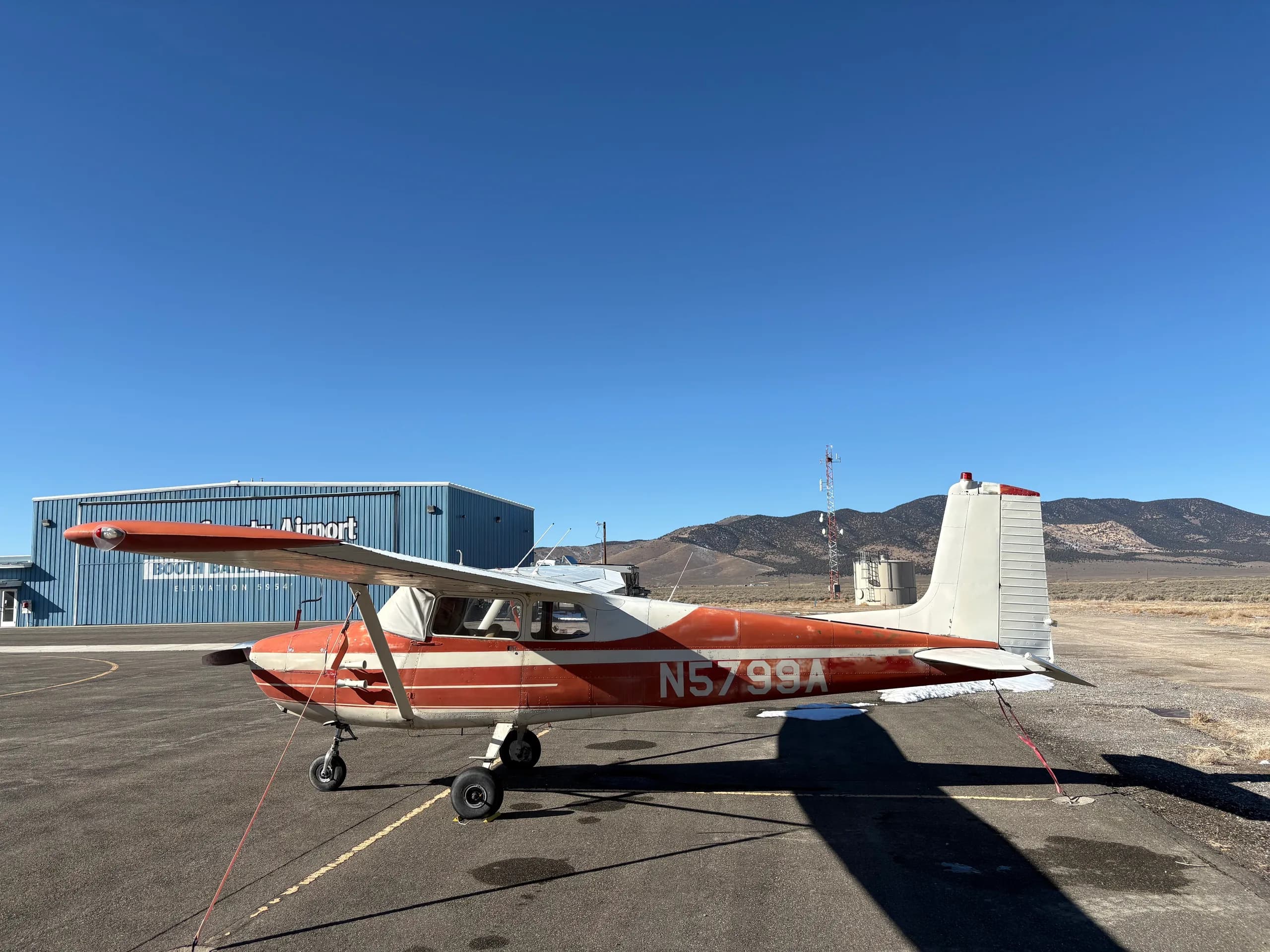 A red and white single-engine high-wing aircraft, N5799A, parked on an airfield with a blue hangar labeled "Eureka County Airport" in the background.