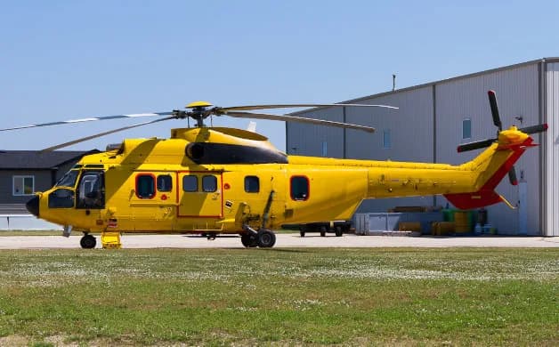 A large yellow helicopter with red tail accents is parked on a grassy airfield next to a hangar under a clear blue sky.