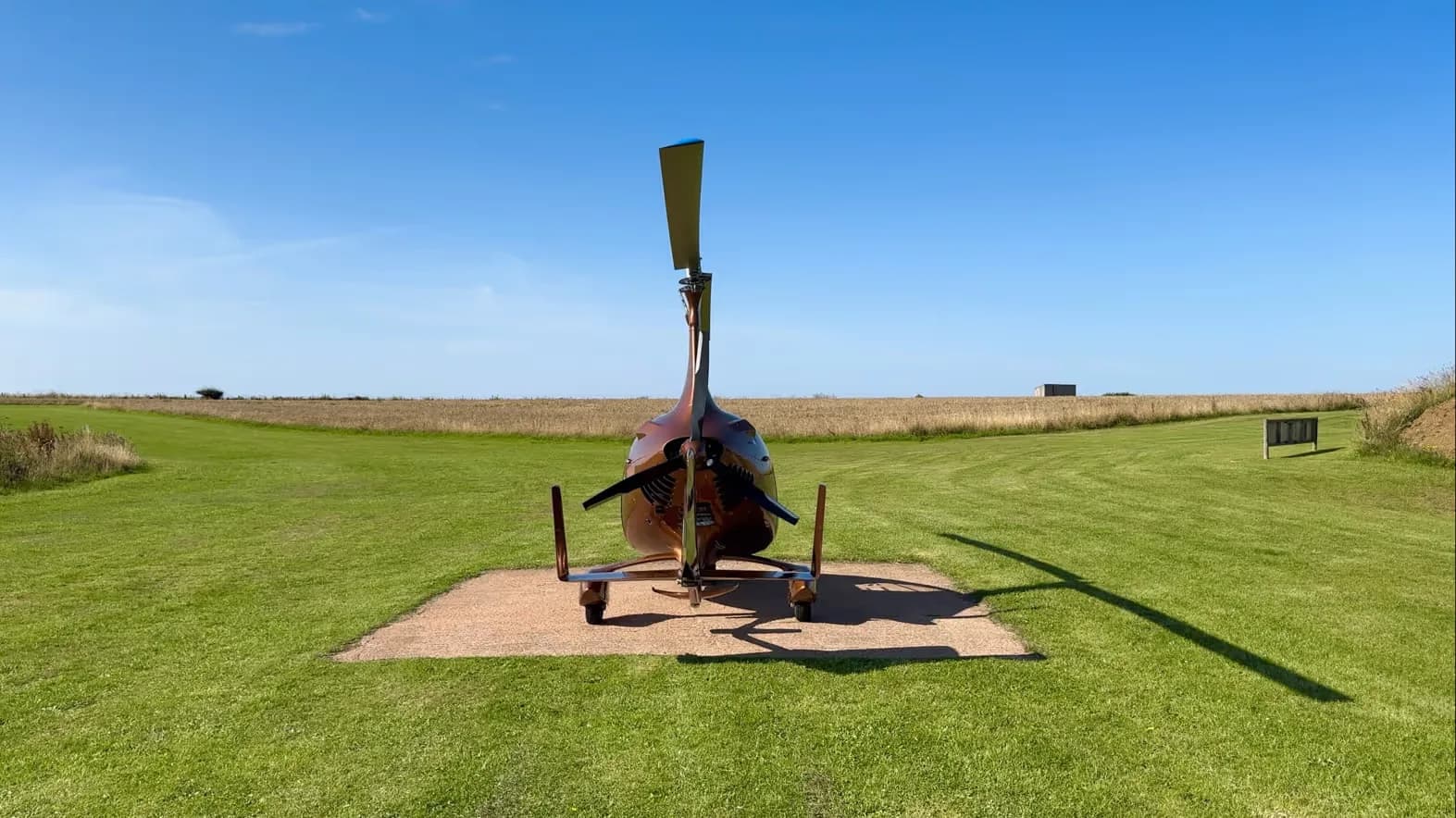 Rear view of a brown gyrocopter parked on a concrete pad in a grassy field.