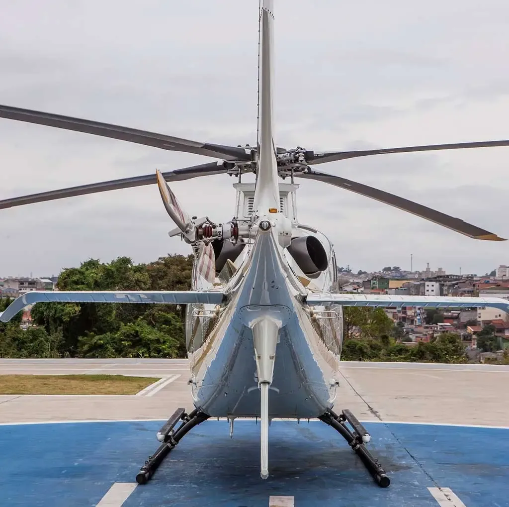 A white helicopter with gold stripes is parked on a blue helipad, viewed from the rear showing the tail rotor.