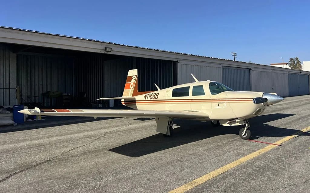 A cream-colored single-engine aircraft with brown and orange stripes, registration N1168B, parked on an asphalt tarmac next to hangars.