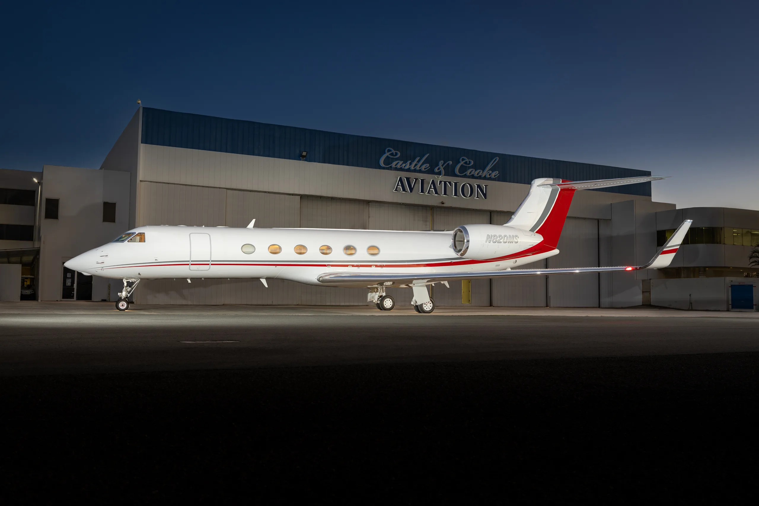 A white Gulfstream G650 with red and gray accents parked on an tarmac in front of a hangar at dusk.