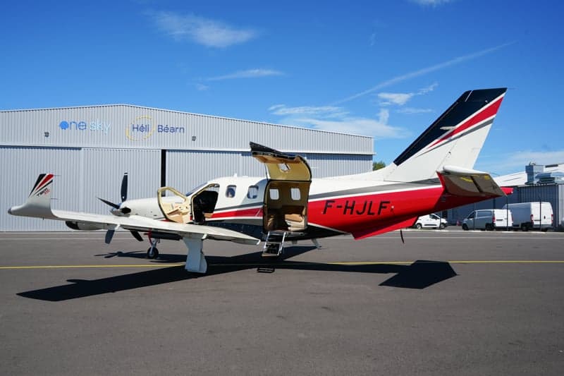 A TBM 930 aircraft with its door open is parked in front of a hangar.