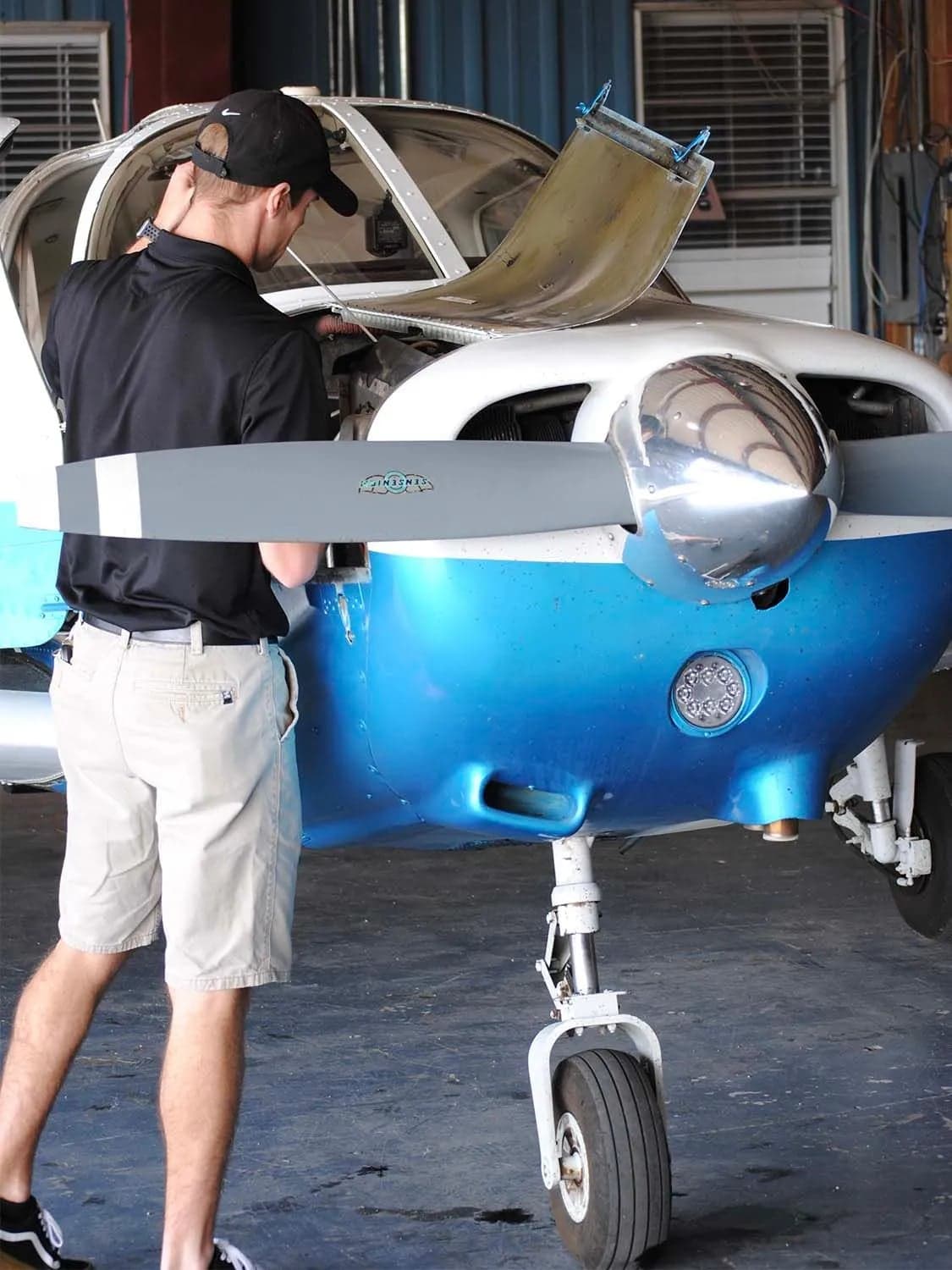 A person in a black shirt and cap works on the engine of a blue and white single-engine aircraft in a hangar.