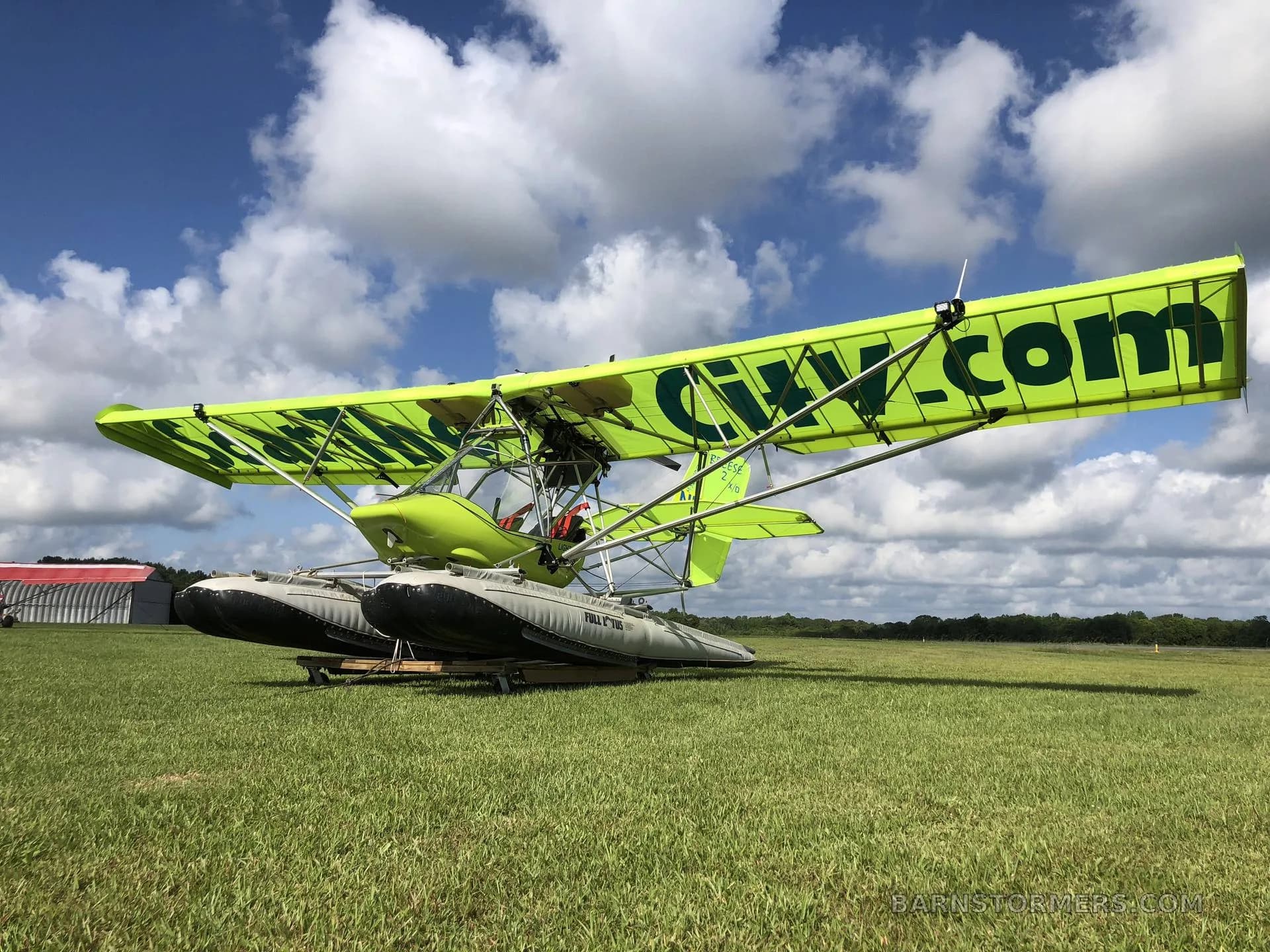 A bright green ultralight seaplane with "Soar The City.com" on its wings is parked on a grassy field under a blue sky with white clouds.